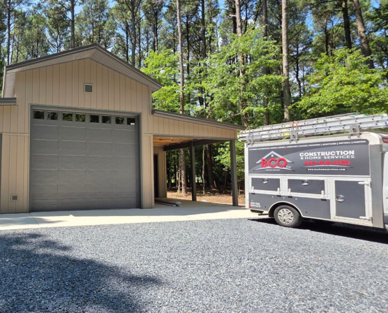 BCO Construction & Home Services trailer parked beside a newly constructed pole barn with a large garage door, covered side area, and wooded surroundings.